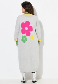 Lange, grijze gebreide cardigan met grote roze, groene en gele bloemmotieven op de achterkant. Beschikt over kleurrijke gestreepte manchetten en kraag.