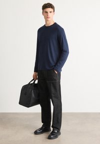 Navy long-sleeve shirt, relaxed fit; black trousers with pockets; black leather bag; black shoes with textured detail; neutral background.