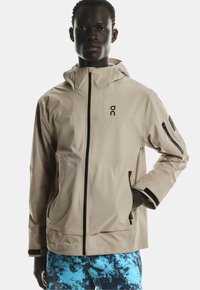 Beige waterdichte jas met zwarte accenten, aansluitende capuchon, met rits afgesloten zakken en een logo op de borst. Soepele textuur, sportieve snit.