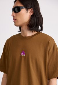 Brun bomull T-skjorte med avslappet passform. Har en lilla ACG-logo brodert på brystet. Modellen har på seg svarte polariserte solbriller.