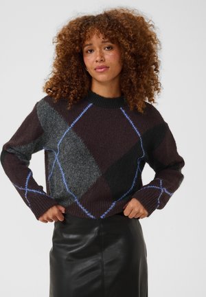 Jeune femme aux cheveux bouclés portant un pull à losanges foncé et une jupe en cuir noire, posant avec les mains sur les hanches devant un fond uni.