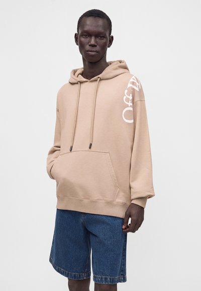 Homme portant un sweat à capuche beige oversize avec un texte blanc sur la manche et un short en jean bleu, debout devant un fond clair uni.