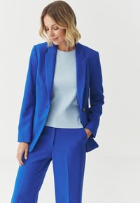 Blue tailored suit jacket and matching pants, paired with a light blue top. Smooth fabric, structured design, and subtle button detail on the jacket.