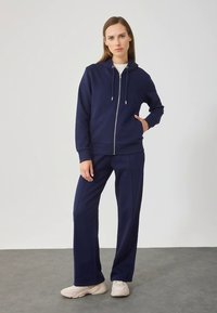 Vrouw staat in een marineblauwe zip-up hoodie en bijpassende broek, met witte sneakers, met één hand in de zak tegen een effen achtergrond.