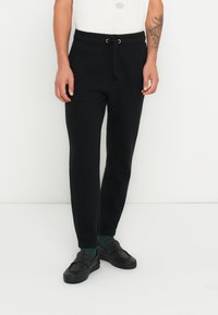 Pantaloni de jogging negri cu talie cu șnur, picioare conice și manșete ribbate, confecționați dintr-un material moale, asortați cu pantofi negri slip-on și șosete verzi.