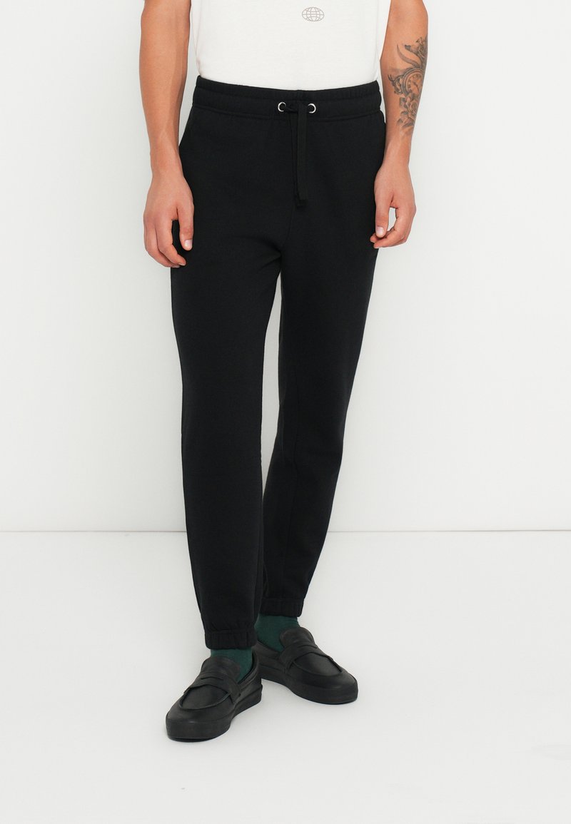 Pantaloni de jogging negri cu talie cu șnur, picioare conice și manșete ribbate, confecționați dintr-un material moale, asortați cu pantofi negri slip-on și șosete verzi.