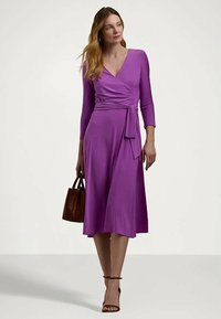 Mulher em vestido roxo com mangas até ao cotovelo, segurando uma carteira castanha, vestindo sapatos de salto com tiras nos tornozelos castanhos, de pé diante de um fundo simples.