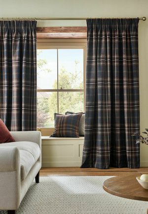 Fenster mit dunkel karierten Vorhängen und passendem Kissen auf beigem Fenstersitz, neben einem hellbeigen Sofa mit rotem Kissen und einem Holzcouchtisch.