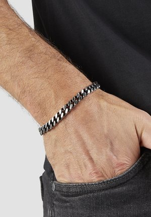 Handgelenk eines Mannes mit einem silbernen Kettenarmband, Hand in der vorderen Tasche einer schwarzen Jeans, trägt ein schwarzes Hemd.