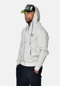 Sweat à capuche zippé gris avec cordons et poches avant, porté avec un pantalon noir. Casquette noire avec des accents colorés et un logo à l'avant.