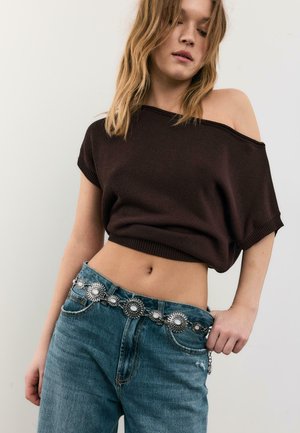 Nainen, joka pukeutuu löysään, tummanruskeaan olkapäät paljastavaan toppiin, korkeavyötäröisiin sinisiin farkkuihin ja koristeelliseen hopeiseen concho-vyöhön, katsoo alas.