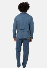 Mies, jolla on kiharaiset hiukset, pukeutuneena siniseen kuviolliseen pitkähihaiseen paitaan ja yksivärisiin sinisiin housuihin, seisoo paljain jaloin selin valkoista taustaa vasten.