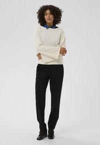 Pull crème texturé avec des manches évasées, porté sur une chemise en denim bleu, associé à un pantalon noir et des chaussures noires brillantes.