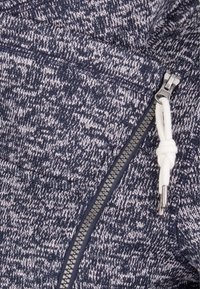 Marinblå stickad huvtröja med en strukturerad mönster, en silverfärgad dragkedja och en snörning med en vit snöre.