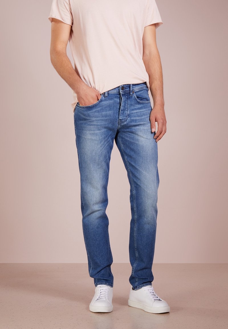 Homme portant un jean slim bleu clair, des baskets blanches et une chemise à manches courtes rose pâle, une main dans la poche.