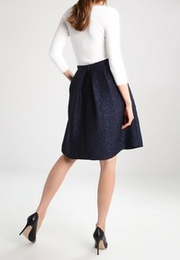 Ein knielanger, marineblauer Rock mit strukturiertem Muster, plissiertem Design und einem Reißverschluss hinten. Trägt dazu ein eng anliegendes weißes Oberteil und schwarze High Heels.