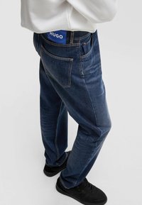 Jeans in denim di colore blu scuro, con una vestibilità regolare, cinque tasche e un'etichetta HUGO ben visibile sulla vita posteriore. Tessuto testurizzato con dettagli di cucitura.