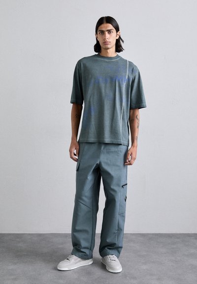 T-shirt vert texturé à manches courtes avec un imprimé graphique, associé à un pantalon cargo teal ample avec poches latérales et fermetures éclair.