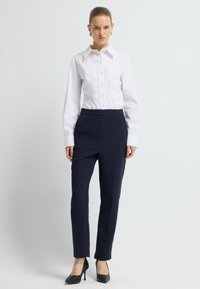 Bílá košile na knoflíky s kapsou, spárovaná s námořnickými kalhotami na míru a černými vysokými podpatky, na neutrálním pozadí.