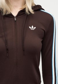 Γυναίκα που φοράει καφέ φούτερ με φερμουάρ, με ανοιχτό γαλάζιες ρίγες στα μανίκια και λευκό λογότυπο Adidas στο στήθος.