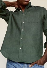 Camisa de lino verde oscuro con mangas largas, tapeta de botones y un bolsillo en el pecho. Textura suave y dobladillo curvado que ofrece un ajuste relajado.
