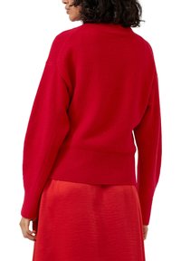 Czerwony dziany sweter z wysokim kołnierzem, opuszczonymi ramionami i prążkowanym dołem, zestawiony z lejącą się czerwoną spódnicą. Widoczne szczegóły tekstury tkaniny.