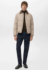 Beżowa pikowana kurtka o gładkiej fakturze, z zatrzaskami i ciemnym kołnierzem, noszona na brązowym swetrze i zestawiona z ciemnoniebieskimi dżinsami.