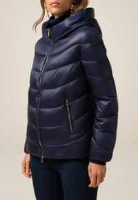 Navyblå dunjakke med hætte, der har vandrette quiltede sektioner, lynlås foran og to sidelommer med metallynlåse.