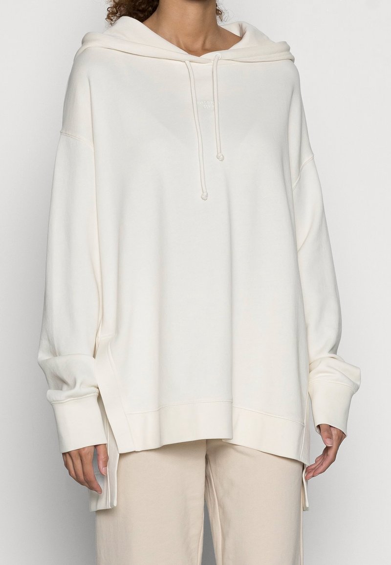 Sudadera con capucha de color crema, hecha de tela suave, con capucha ajustable con cordón, hombros caídos y aberturas laterales para un ajuste holgado.