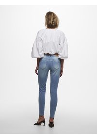 Jeans skinnies azul claro con efecto desgastado y bolsillos traseros, combinados con una blusa blanca de mangas abullonadas. Unas sandalias de tacón negras completan el look.