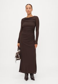 Brun langermet kjole med rynkedetaljer, tettsittende silhuett og slank kutt. Modellen har på seg elegante ankelstøvletter og holder en liten veske.