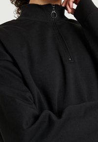 Pullover nero a zip parziale realizzato in tessuto morbido. Presenta un colletto a coste, cuciture laterali e una cerniera metallica con un tirante rotondo.