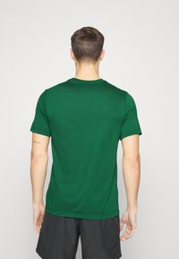 Hombre de pie de espaldas a la cámara, vistiendo una camiseta verde y pantalones cortos negros, mostrando un tatuaje de "mantente salvaje" en el antebrazo izquierdo.