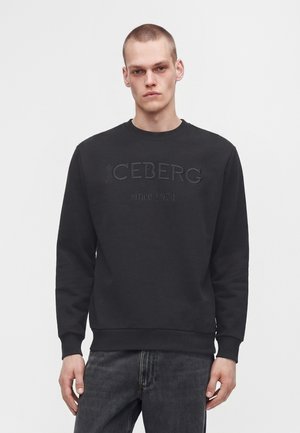 Jeune homme aux cheveux courts portant un sweat-shirt noir avec "ICEBERG since 1974" embossé à l’avant et un jean gris foncé.