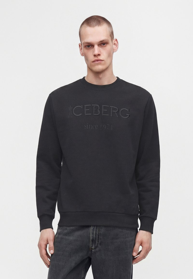 Jeune homme aux cheveux courts portant un sweat-shirt noir avec "ICEBERG since 1974" embossé à l’avant et un jean gris foncé.