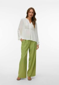 Femme debout, une main dans la poche, portant un chemisier blanc avec des panneaux en dentelle, un pantalon vert à jambes larges et des sandales à brides sur fond blanc.