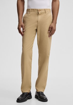 Uomo con pantaloni beige a gamba dritta, camicia bianca e mocassini in pelle neri, in piedi davanti a uno sfondo grigio neutro.
