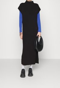 Schwarzes, geripptes Strickkleid mit hohem Kragen, über ein blaues Langarmshirt getragen. Kombiniert mit schwarzen klobigen Schuhen und einer schwarzen Handtasche.