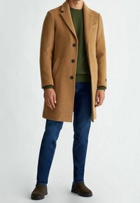 Manteau en laine camel avec revers crantés, trois boutons et poches latérales, associé à un pull vert, un jean bleu et des chaussures marron à enfiler.