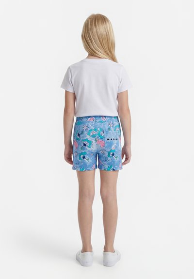 Vaikas su ilgais šviesiais plaukais, dėvintis baltą marškinėlius, mėlynas gėlių rašto šortus su „Marni“ logotipu ir baltus sportbačius, stovintis nugarą atsukęs baltame fone.