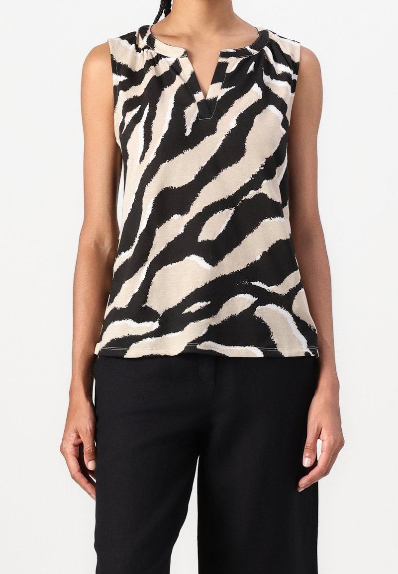 Top sans manches avec des rayures zébrées abstraites beige, noir et blanc, encolure en V entaillée, porté avec un pantalon noir par une personne debout.