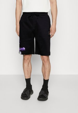 Homme portant un short noir à cordon avec un détail floral violet sur la jambe gauche, des chaussettes noires et des sandales noires sur fond blanc.