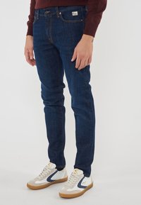 Jeans blu scuro con una vestibilità slim, caratterizzati da un classico design a cinque tasche, cuciture arancioni e una piccola etichetta con logo sulla vita.
