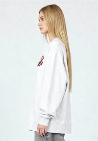 Sweat-shirt gris clair oversize à manches longues, poignets côtelés, col rond, avec un logo brodé rouge et noir sur le devant.