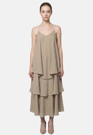 Donna che indossa un vestito beige senza maniche a strati con spalline sottili e tacchi alti beige abbinati, in piedi su uno sfondo semplice.