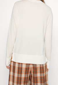 Svetle krémový pletený svetr s kulatým výstřihem, dlouhými rukávy a bočními rozparky na spodním lemu, spárovaný s širokými kalhotami s kostkovaným vzorem v hnědé a oranžové barvě.