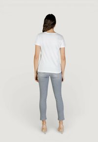 Femme aux longs cheveux bruns portant un t-shirt blanc à manches courtes, un jean gris clair ajusté et des chaussures à talons beiges, debout, de dos.