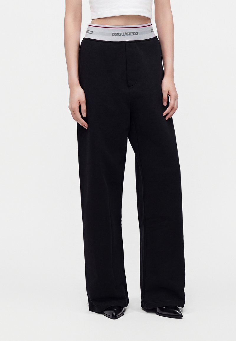 Personne portant un pantalon noir à jambes larges avec une ceinture élastique grise portant l'inscription DSQUARED2 et un top court blanc.