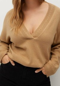 Beige neulepusero, jossa on V-pääntie ja ribbireunus. Muotoilu sisältää pitkät hihat ja rento istuvuus, käytetty mustien housujen päällä.