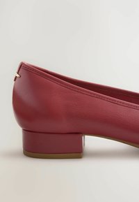 Bailarina de cuero rojo con un bajo tacón cuadrado, que presenta un borde cosido y un pequeño acento dorado en la parte posterior. Textura suave.